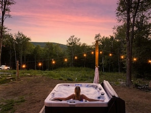 A person relaxing in a hot tub outdoors with string lights in a forested area.