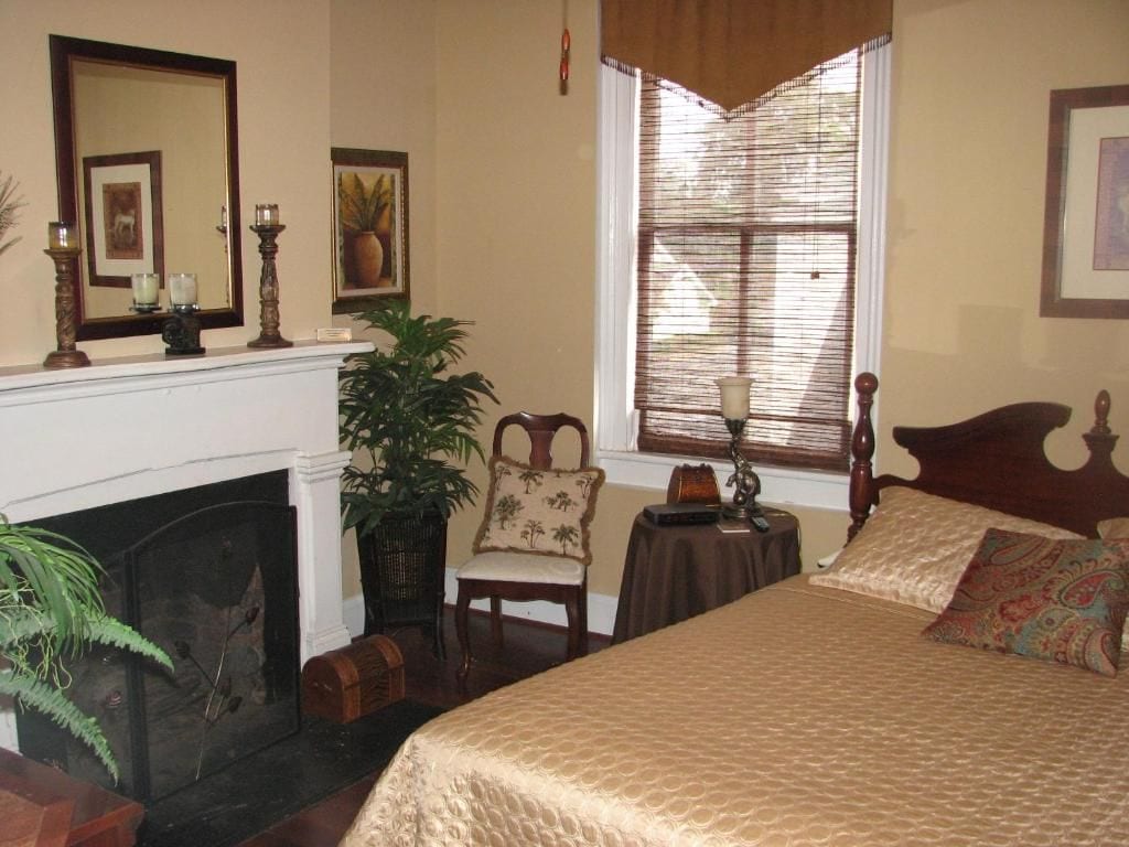 A bedroom with a fireplace, a bed with patterned bedding, a chair, a small table, and a window with blinds.