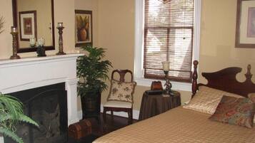 A bedroom with a fireplace, a bed with patterned bedding, a chair, a small table, and a window with blinds.