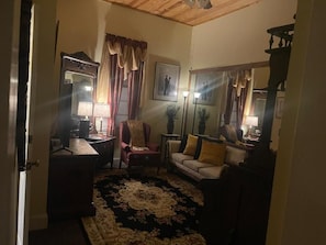 A room with a wooden ceiling, a sofa, a chair, a small table, a lamp, a mirror, and a framed picture on the wall.