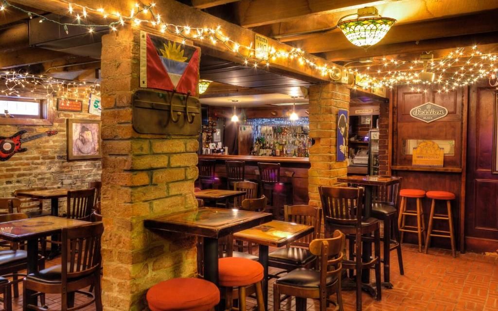An indoor bar with brick walls, wooden tables, and chairs. There are string lights, a flag, and a sign on the wall.
