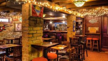 An indoor bar with brick walls, wooden tables, and chairs. There are string lights, a flag, and a sign on the wall.