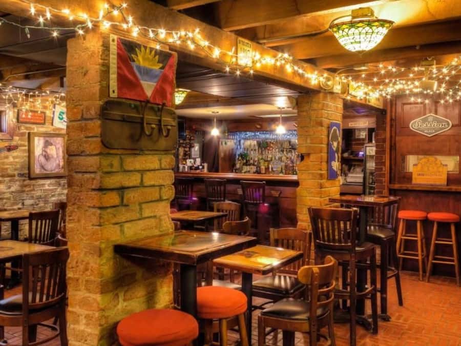 An indoor bar with brick walls, wooden tables, and chairs. There are string lights, a flag, and a sign on the wall.