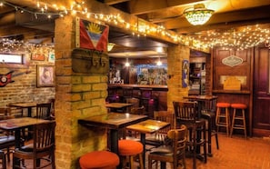 An indoor bar with brick walls, wooden tables, and chairs. There are string lights, a flag, and a sign on the wall.