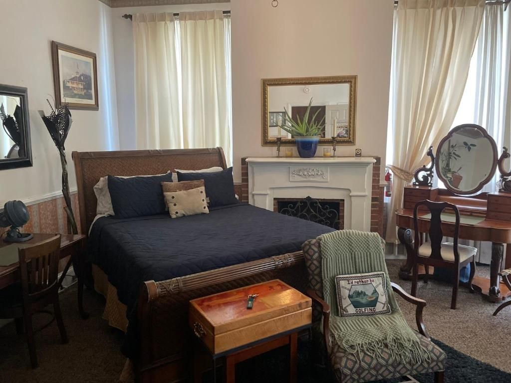 A bedroom with a bed, a dresser, a chair, a fireplace, and a mirror.