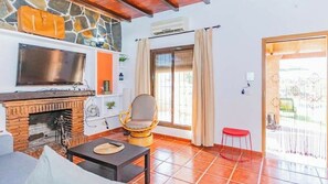 Uma sala de estar com lareira de pedra, televisão, cadeira de vime e um banquinho vermelho.