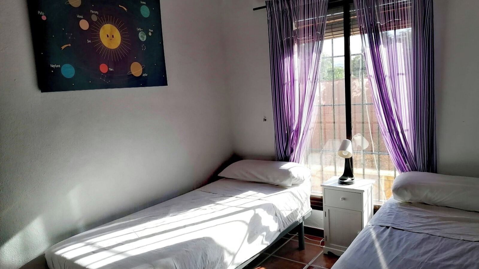 A bedroom with two single beds, a nightstand with a lamp, a window with purple curtains, and a wall-mounted poster of the solar system.