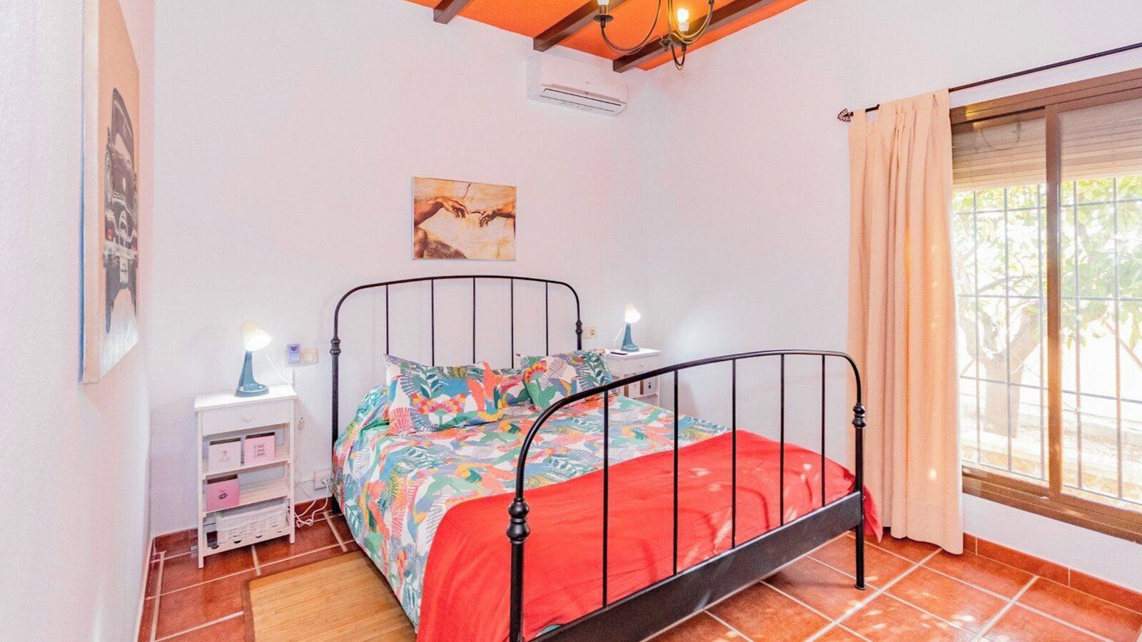 Ein Schlafzimmer mit Bett, Nachttisch, einem Fenster mit Vorhängen und einem Wandbild.