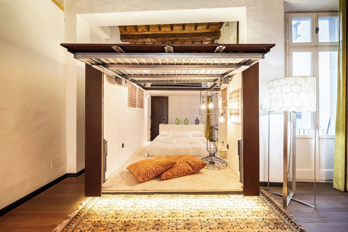 A bedroom with a bed, a floor lamp, and a view of the outside.