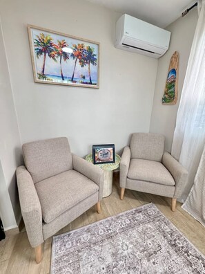 A living room with two armchairs, a small round table, a framed picture of a beach scene, an air conditioning unit, and a patterned rug. - Beautiful 3-bedroom house with ocean view and amazing sunsets in charming Rincon (Rincon)