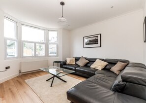 A modern living room with a leather sofa, a glass coffee table, and a large window with blinds. - Luxury 4BR Kingston | London | Villa | Garden Oasis | Free Parking | Near Train (Kingston upon Thames)