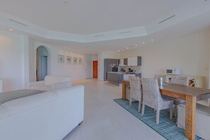 A modern living room with a white sofa, a dining table with wicker chairs, and a kitchen in the background. - Love Beach E2 - Luxury 3BR Condo + Pool & Beach (Nassau)