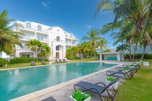A resort with a swimming pool, lounge chairs, palm trees, and a white building with balconies. - Love Beach E2 - Luxury 3BR Condo + Pool & Beach (Nassau)