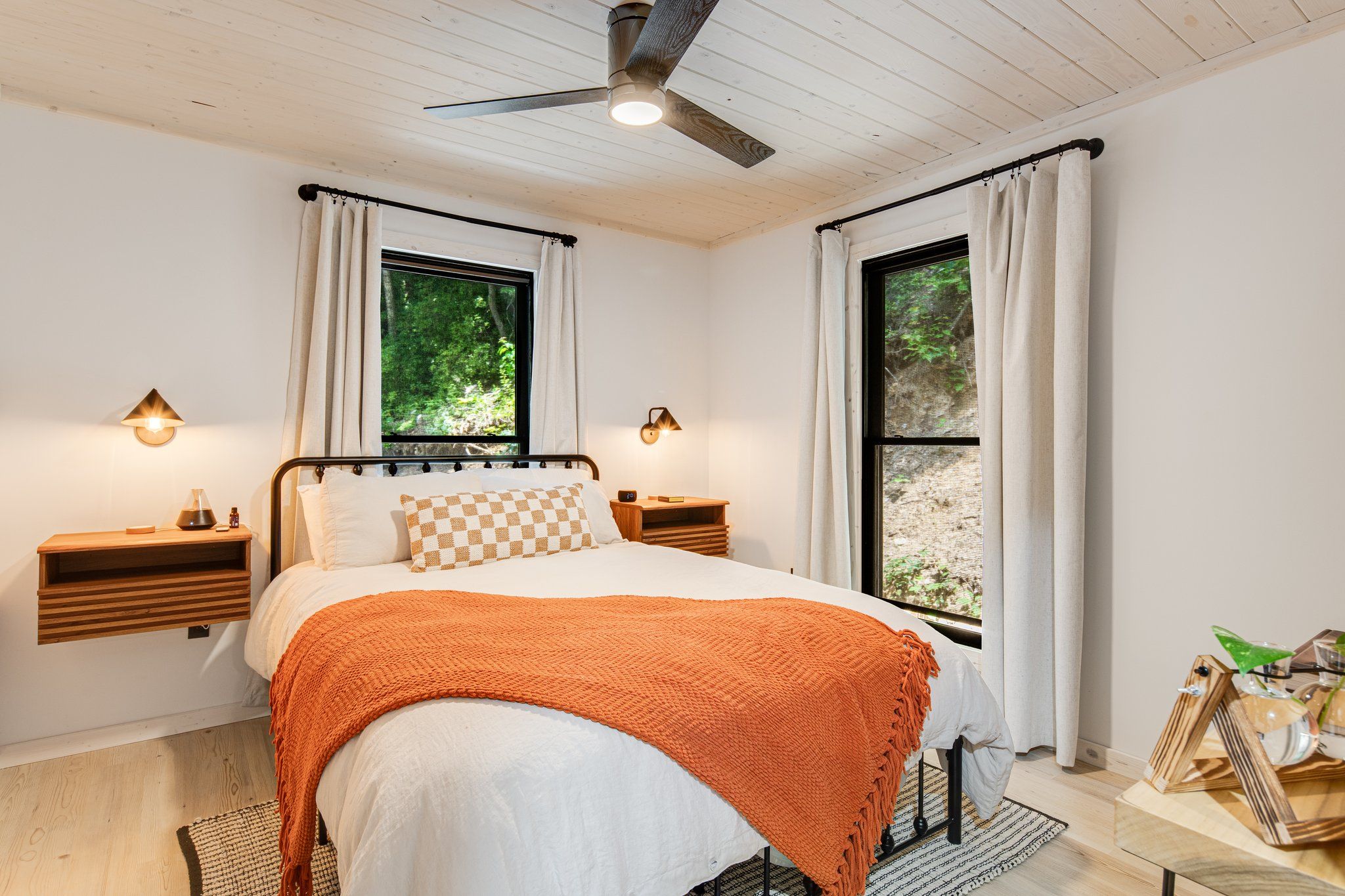 Quarto com cama, duas mesas de cabeceira, ventoinha no teto e janelas com cortinas.