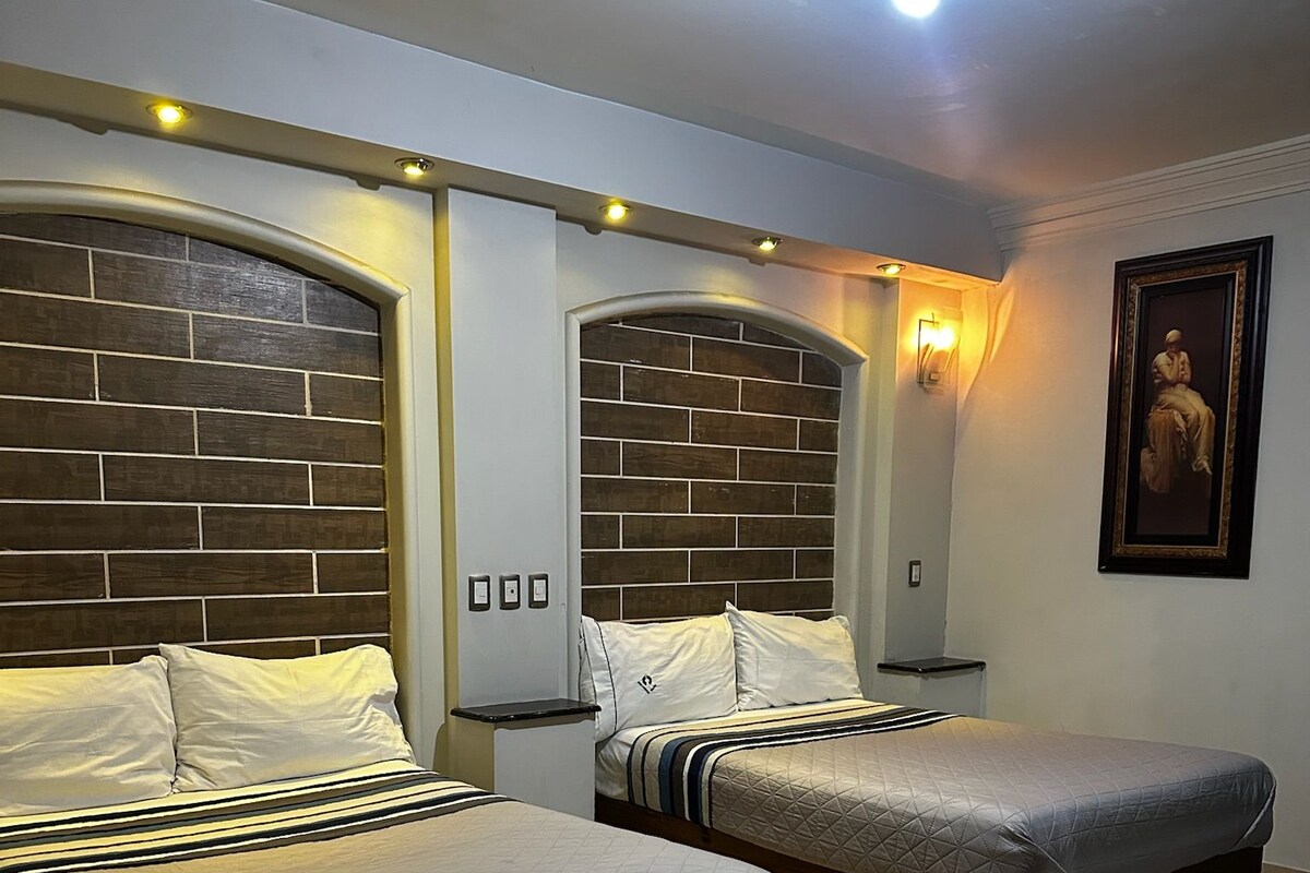 A hotel room with two beds, a brick-patterned headboard, a nightstand, a wall-mounted light, and a framed picture.
