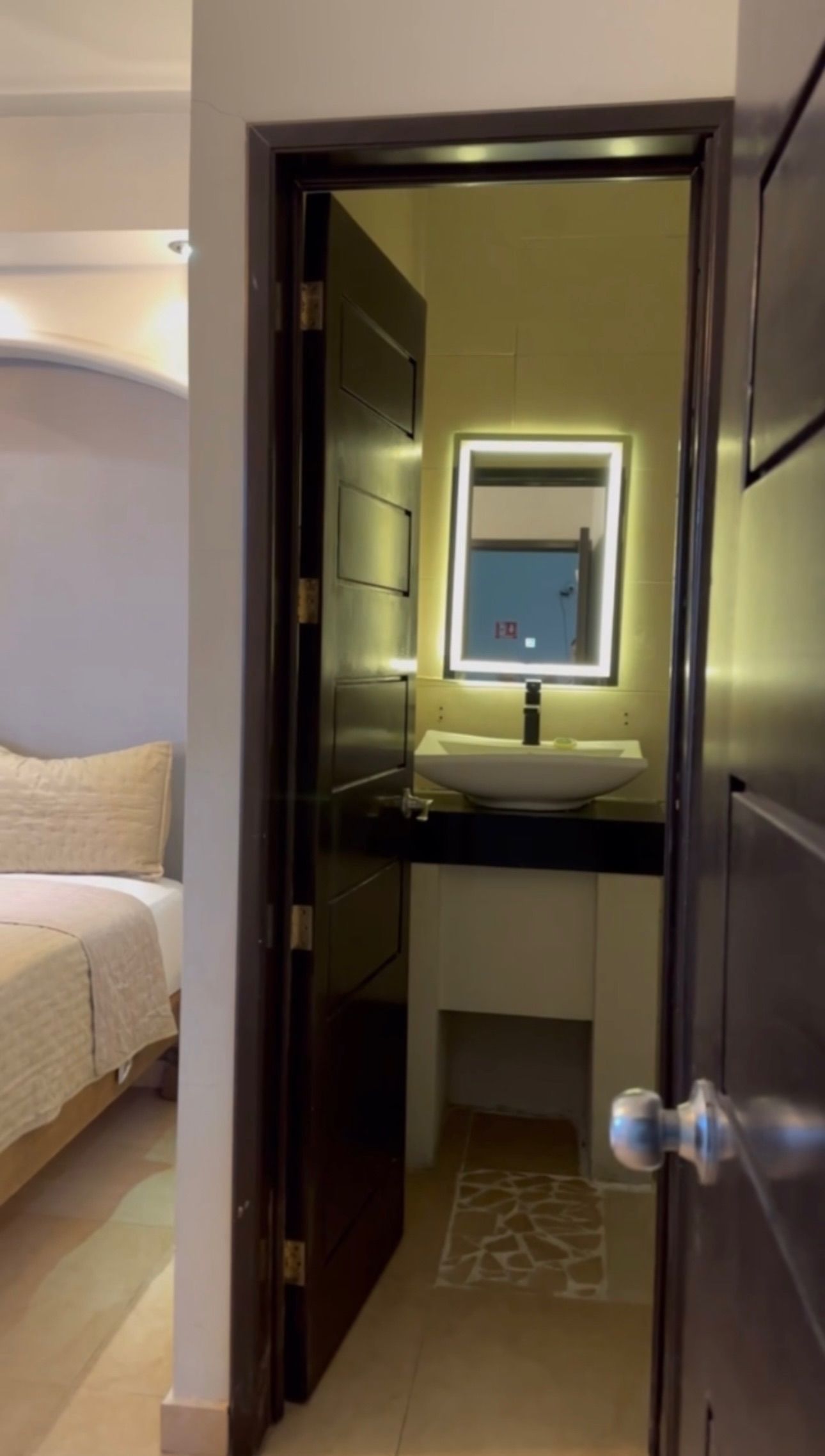 Una habitación de hotel con una cama, un baño con un espejo y un lavamanos, y una puerta que da a otra habitación.
