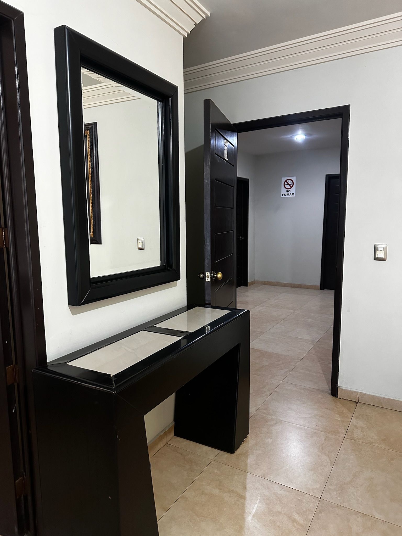 Un pasillo con una consola negra, un espejo y una puerta con el letrero 'NO FUMAR'.