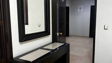 Un pasillo con una consola negra, un espejo y una puerta con el letrero 'NO FUMAR'.