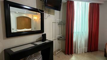 Habitación de hotel con televisor de pantalla plana montado sobre una consola negra, un espejo grande, un ventilador, cabina de ducha de vidrio y cortinas rojas.