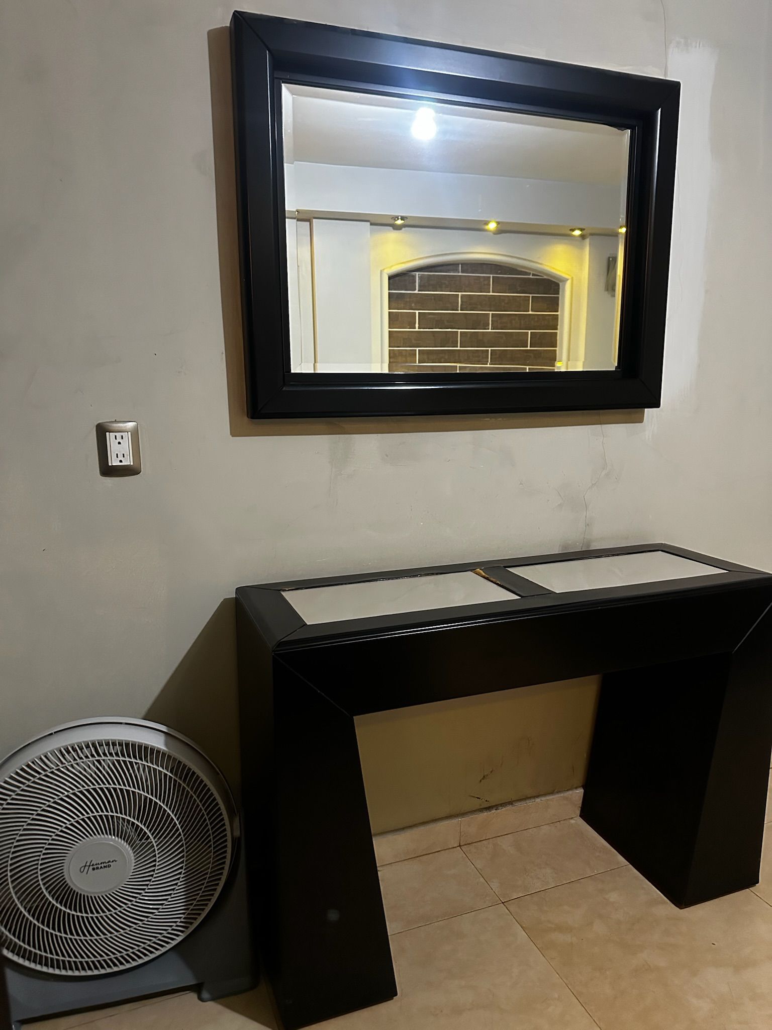 A room with a black-framed mirror, a black desk with a white top, and a standing fan.