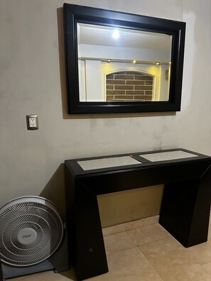 Una habitación con un espejo de marco negro, un escritorio negro con superficie blanca y un ventilador de pie.