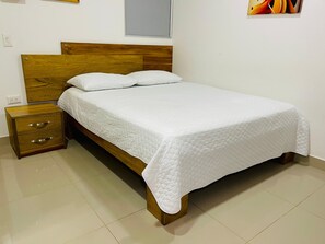 Uma cama de solteiro com cabeceira e peseira de madeira, uma pequena mesa de cabeceira com gavetas, e uma parede simples com uma janela sobre a cama.