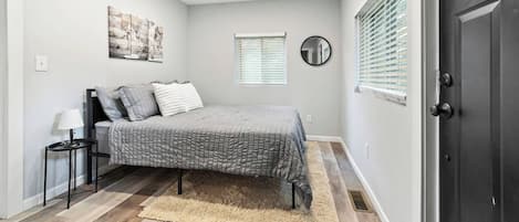 A bedroom with a bed, a bedside table, a lamp, a rug, a window with blinds, and a door.