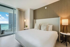 Habitación de hotel con una cama grande, vista al océano, una mesita con lámpara y una ventana con cortinas.