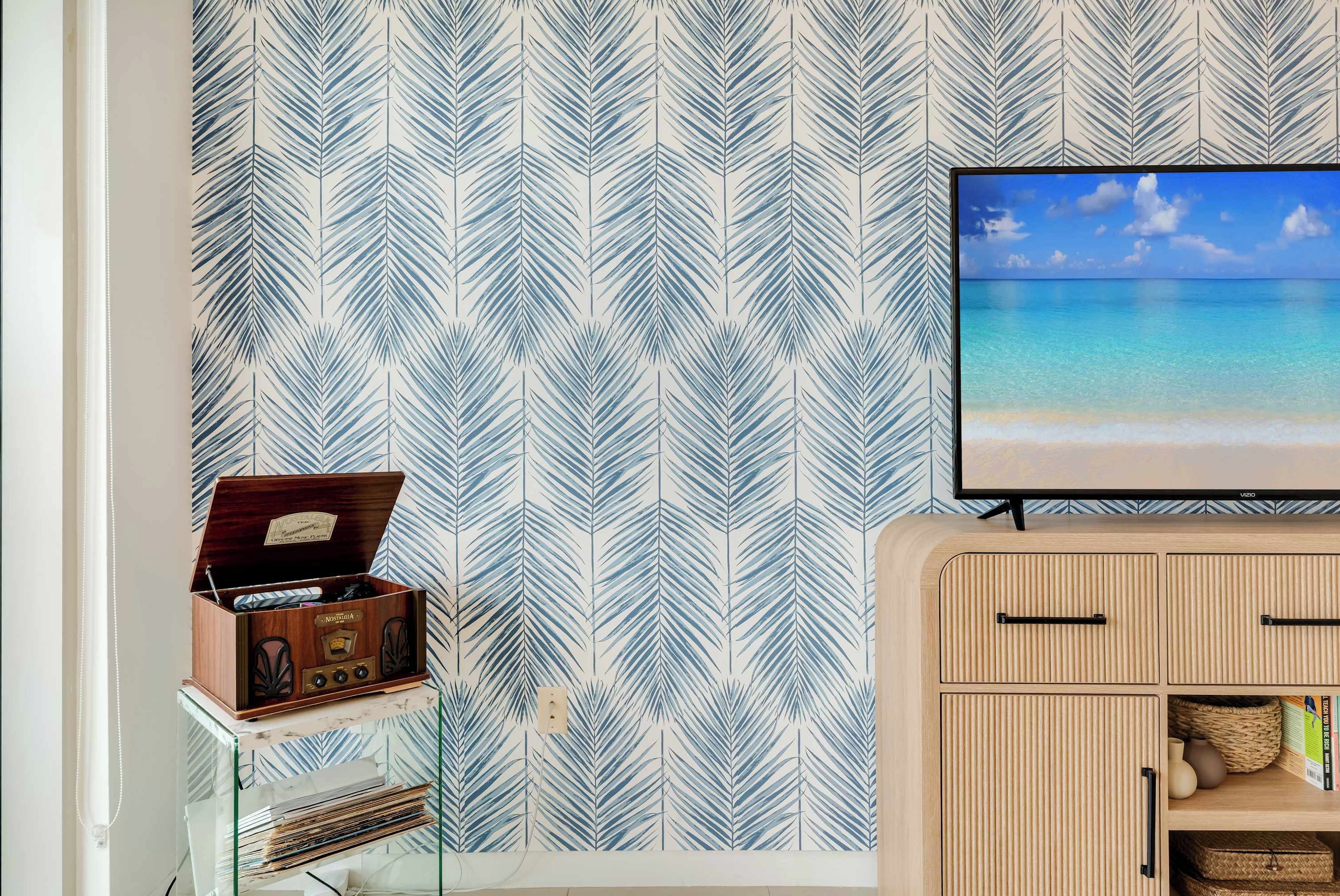 A room with a patterned wallpaper, a wooden radio, a television on a stand, and a wooden cabinet with drawers.
