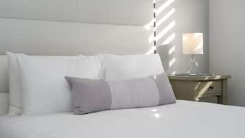Una cama bien tendida con ropa de cama blanca y gris, una mesita de noche con una lámpara, y una pared con un ventanal que proyecta sombras.