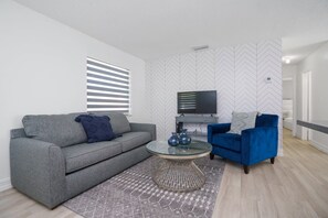 A modern living room with a grey sofa, a blue armchair, a glass coffee table, and a TV on a white wall with a chevron pattern.