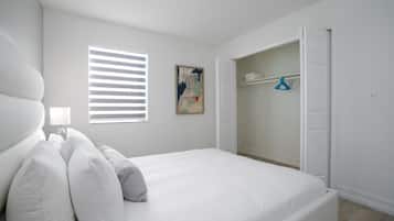 A bedroom with a bed, a window with blinds, a painting, and a closet with hanging clothes.