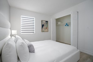 A bedroom with a bed, a window with blinds, a painting, and a closet with hanging clothes.