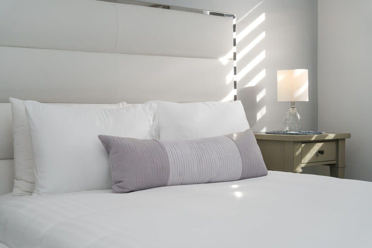 Una cama bien hecha con cabecera, una mesita de noche con lámpara, y una pared con ventana que proyecta sombras.