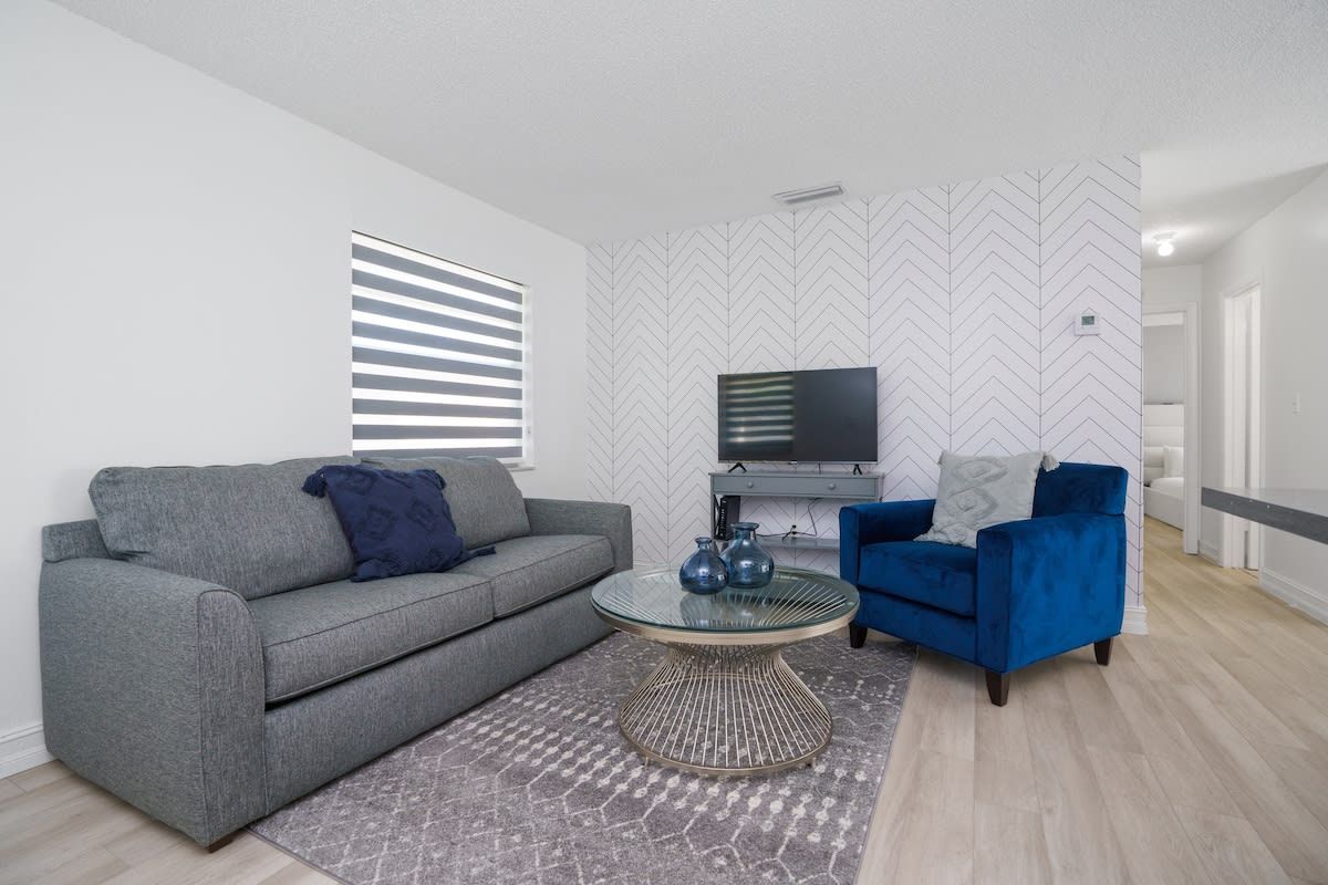 Uma sala de estar moderna com um sofá cinzento, uma poltrona azul, uma mesa de centro de vidro e um tapete estampado.