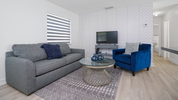 A modern living room with a grey sofa, a blue armchair, a glass coffee table, and a patterned rug. - Cozy Blue Door Duplex, Central in Cutler Bay (Cutler Bay)