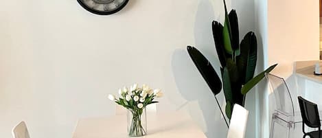 A dining area with a white table, four chairs, a vase with flowers, a wall clock, and a large plant.
