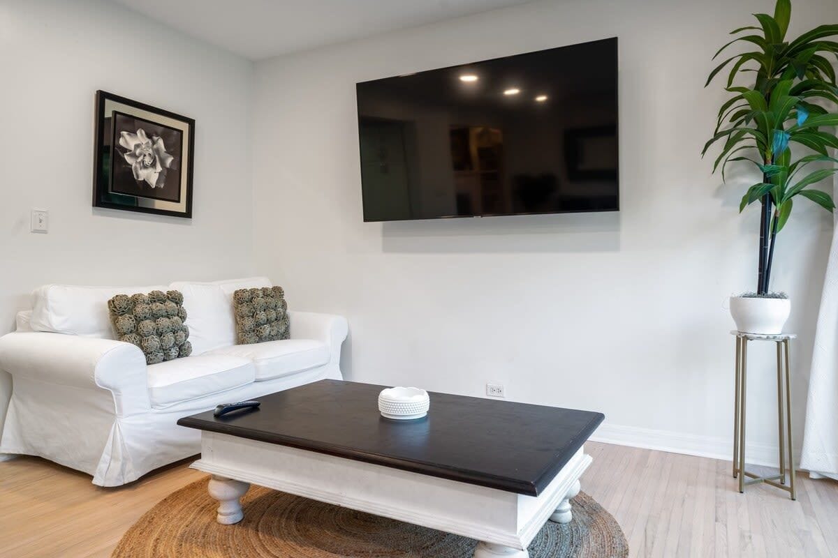 Uma sala de estar moderna com um sofá branco, uma mesa de centro escura, uma televisão de tela plana e um vaso com planta.