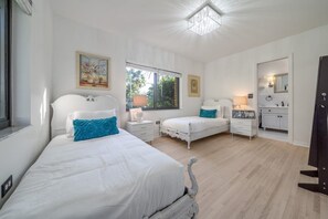 Un dormitorio con dos camas, un ventanal grande, una lámpara de araña y un baño visible a través de una puerta abierta.