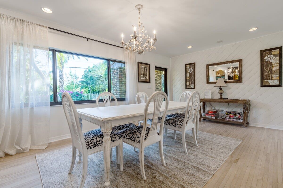 A dining area with a chandelier, white chairs with patterned cushions, a white table, and a view of greenery outside.