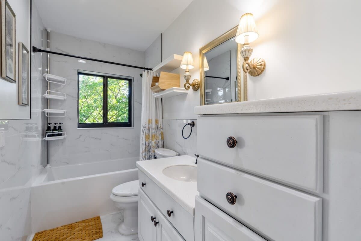 A modern bathroom with a bathtub, toilet, vanity with drawers, and a mirror with wall sconces.