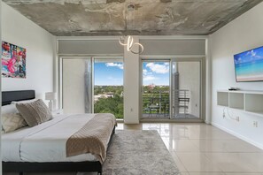 Un dormitorio moderno con cama, un ventanal grande, balcón y vista a la ciudad.