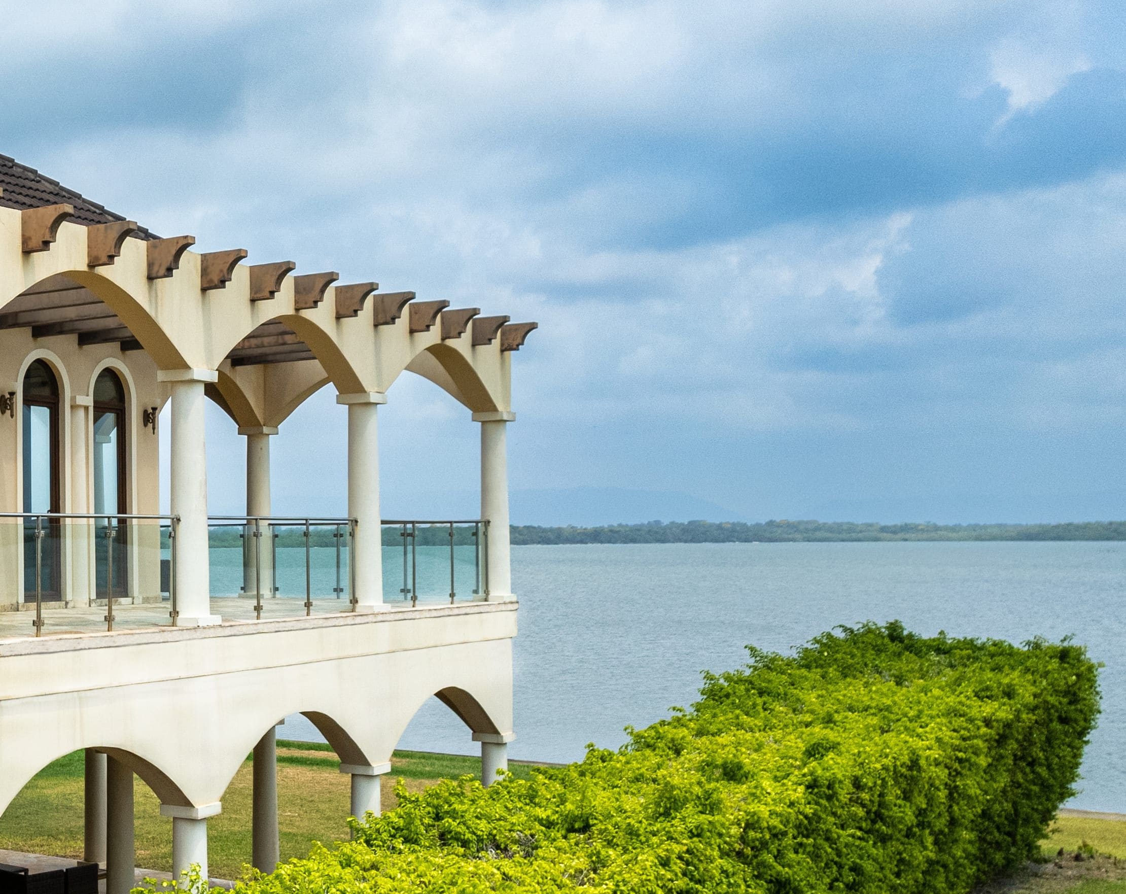 A building with a balcony overlooking a body of water, supported by columns, with a clear sky above.