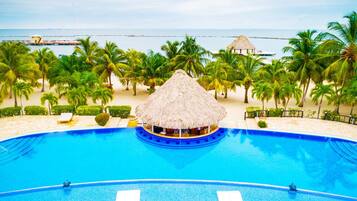 A large, clear blue pool with a thatched-roof bar area, surrounded by palm trees and a beach view.