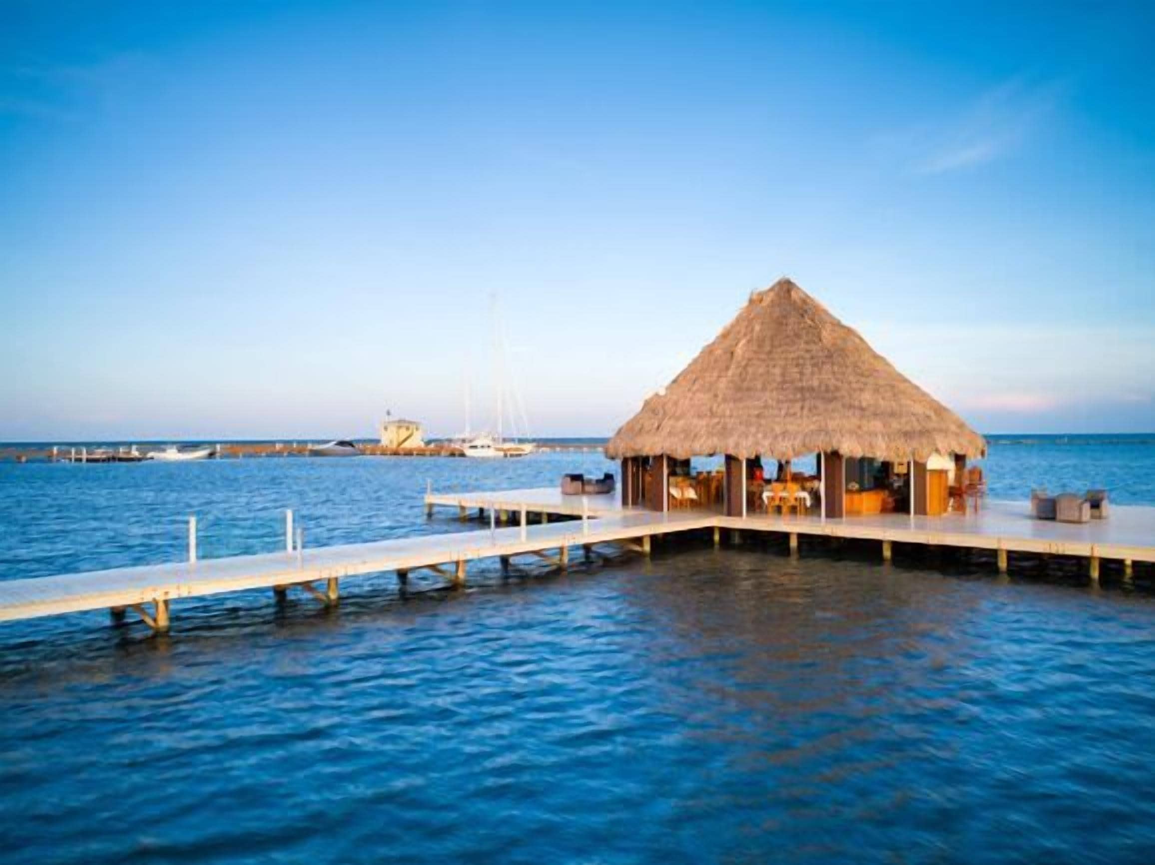 A thatched-roof overwater bungalow with a wooden deck extending into the water, a clear sky, and distant boats.