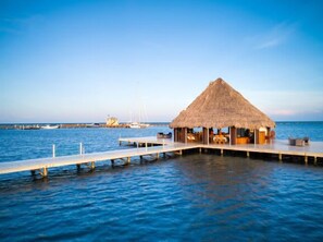A thatched-roof overwater bungalow with a wooden deck extending into the water, a clear sky, and distant boats.