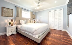 Un dormitorio con cama, mesitas de noche, ventilador de techo, ventana con cortinas, espejo montado en la pared y armario.