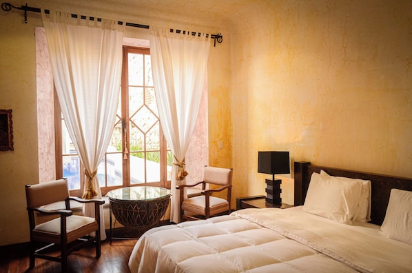 A hotel room with a bed, two chairs, a small table, a lamp, and a window with curtains. - Boutique Magia Stay, Pool in Antigua (Antigua Guatemala)