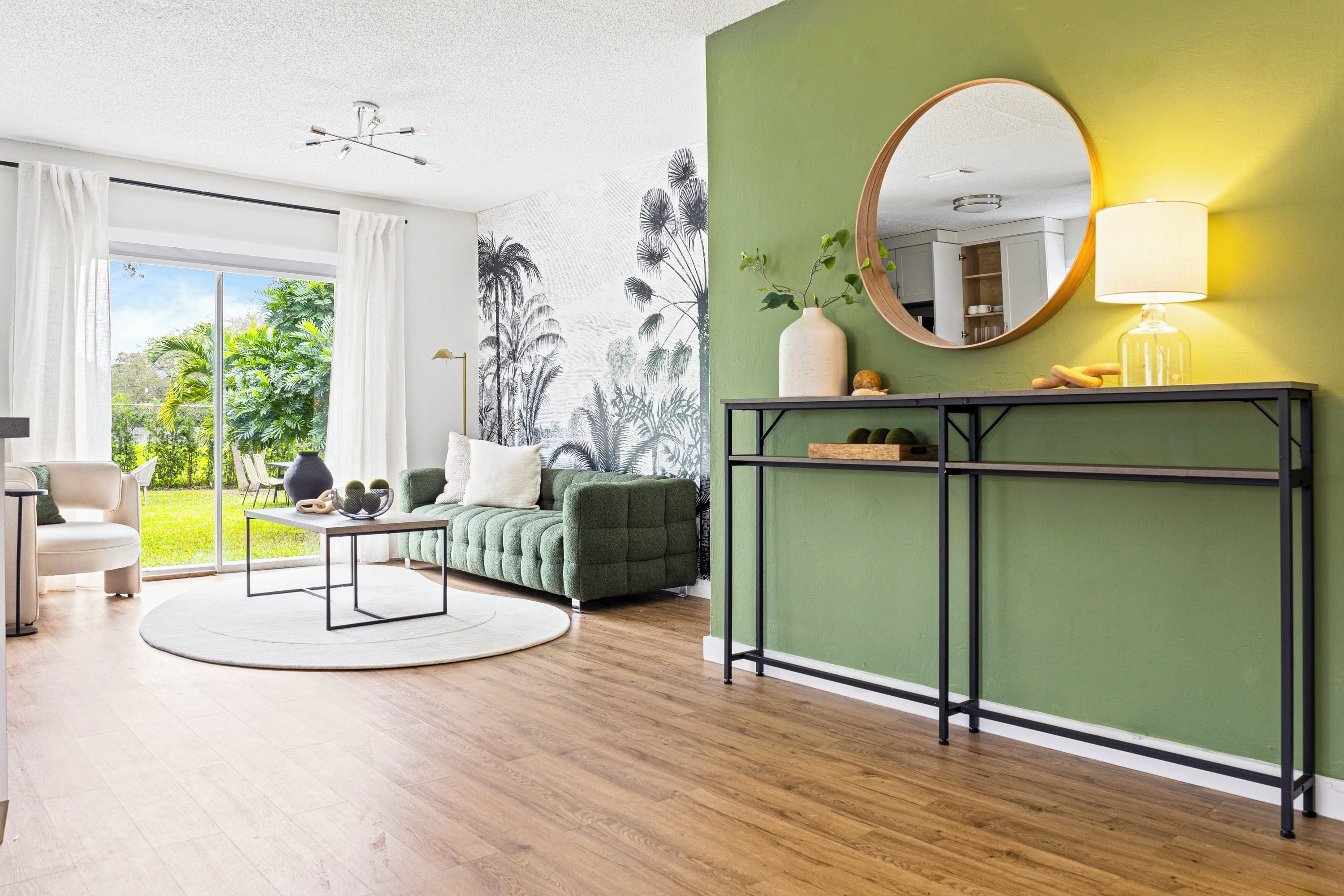 Uma sala de estar moderna com uma parede verde de destaque, um espelho redondo, um aparador e vista através de uma porta de vidro.