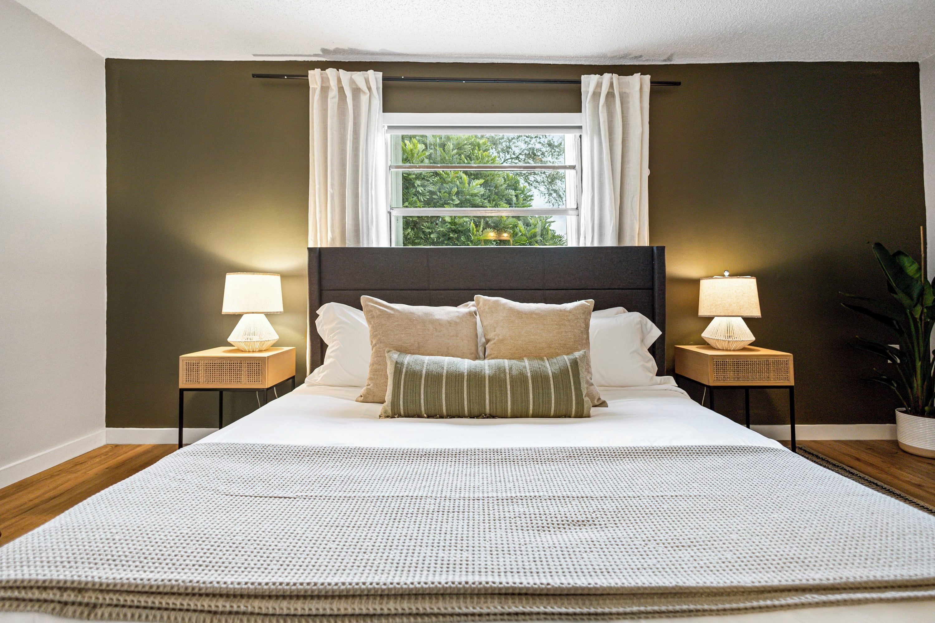 Une chambre avec un grand lit, deux tables de chevet avec des lampes, une fenêtre avec des rideaux et une vue sur la verdure à l’extérieur.